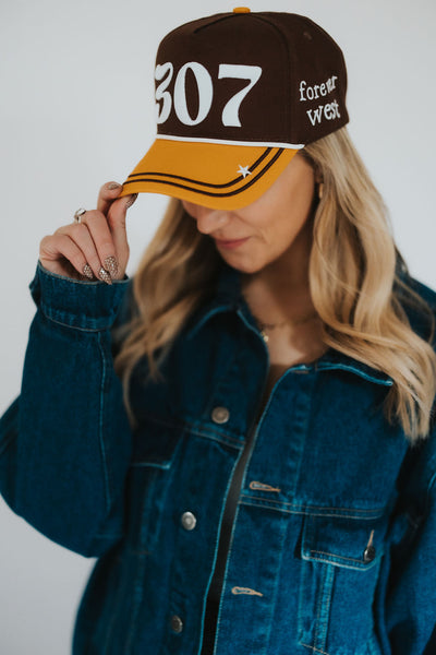 Woman in a denim jacket grabs the brim of her RUBY WYLD 307 Forever West brown and gold hat 