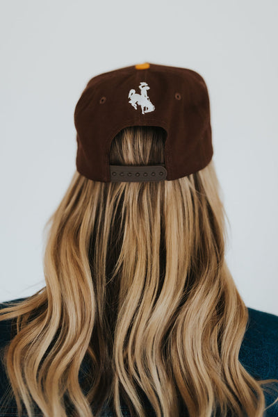 Back of woman's head wearing a brown and gold Cowboys hat with a white bucking horse over the snaps 