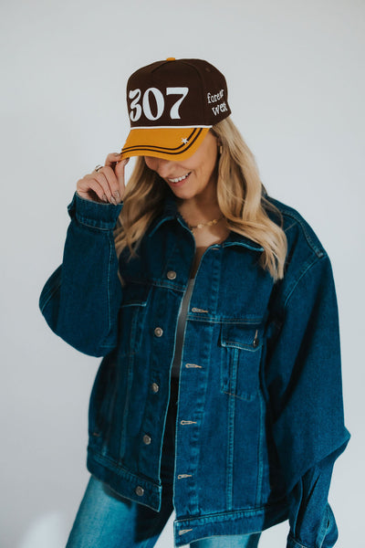 Wyoming cowgirl poses in a denim western jacket as she smiles and grabs the brim of her Wyoming Cowboys brown and gold snapback hat 