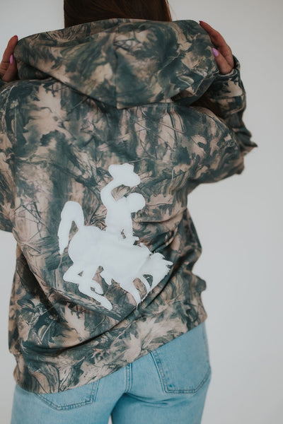 woman puts a zip-up sweatshirt hood on her head exposing the large, white bucking horse University of Wyoming logo embroidered on the back of the came hoodie 