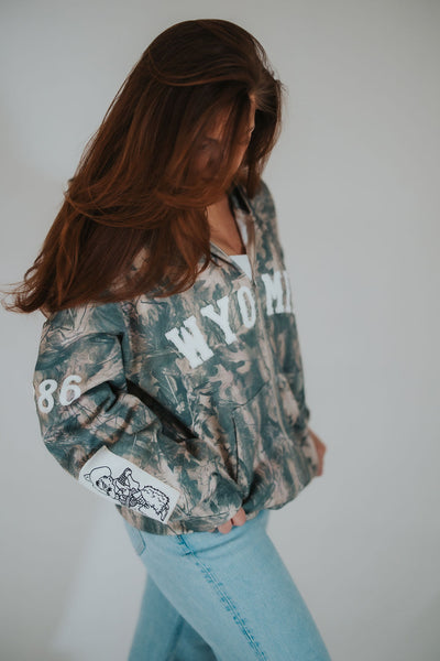 Woman looks down and poses in RUBY WYLD's custom, University of Wyoming licensed camo Wyoming hoodie with embroidered Pistol Pete on the sleeve