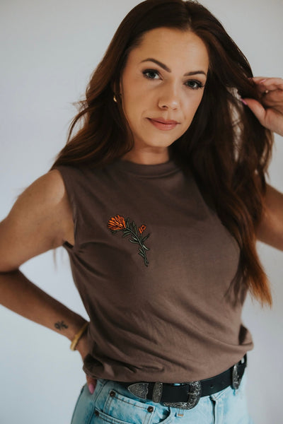 Fashionable Wyoming woman brushes hair back as she poses in RUBY WYLD's muscle tank tee in brown with an embroidered indian paintbrush on the right chest 