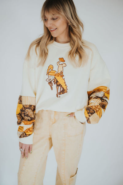 Woman poses in a women's cream crewneck sweater with patchwork sleeves and the Wyoming Cowboys bucking horse on the front 