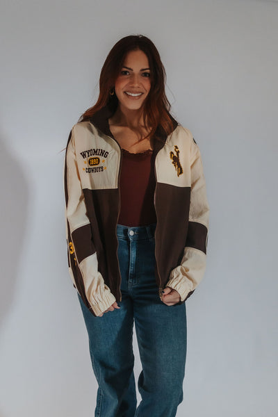 A woman wearing a color-blocked vintage-style windbreaker jacket with cream and brown panels, featuring 'Wyoming Cowboys' text and a bucking horse logo on the chest, paired with a dark brown top and blue jeans, against a plain light gray background.