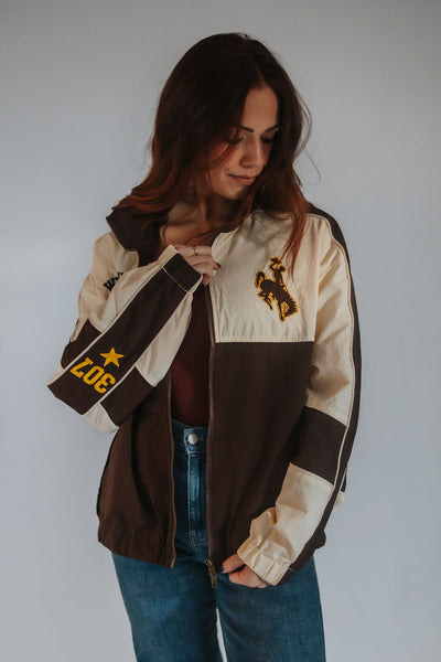 A woman wearing a vintage-style colorblock windbreaker jacket in brown and cream, featuring a bucking horse patch on the chest and '307' with a star on the sleeve, paired with blue jeans against a plain gray background.