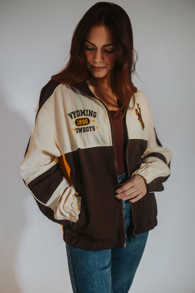 A woman wearing a Wyoming Cowboys varsity jacket in brown and cream, styled with a dark brown top and blue jeans against a plain light gray background.