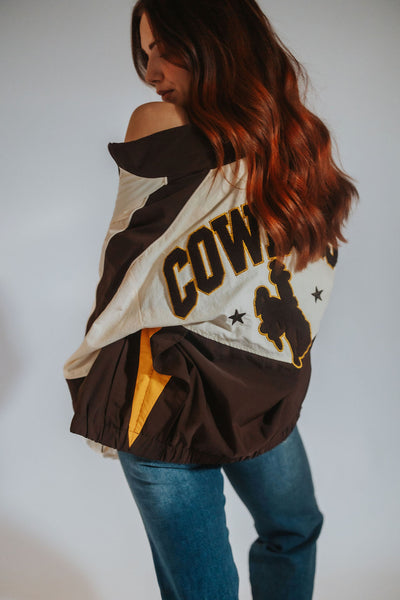 A woman wearing a brown and cream windbreaker jacket with 'COWBOYS' and a bucking horse and rider silhouette graphic, paired with blue jeans, shown from a side-back angle against a plain light background.