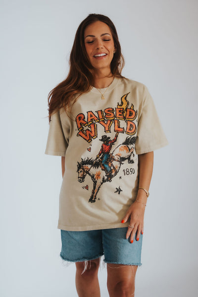 A woman wearing a beige graphic t-shirt with a western theme, featuring the text 'RAISED WYLD 1890' and an illustration of a cowboy riding a bucking horse. 