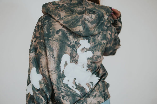 Woman pulls up hood on RUBY WYLD's story-led, University of Wyoming licensed oversized camo zip-up sweater with the Steamboat bucking horse embroidered on the back in white