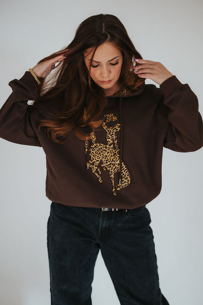 Woman brushes her hair back and looks down at her exclusively designed RUBY WYLD brown hoodie with cheetah print steamboat  bucking horse on the front 