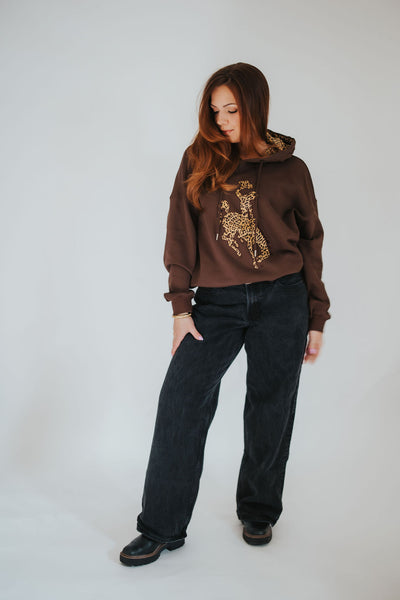 Woman leans into her hip and looks over her shoulder in her University of Wyoming steamboat hoodie in brown with animal print detailing