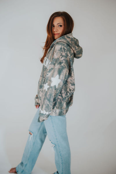 red head woman peeks over her shoulder as she wears an oversized, worn in zip-up Wyoming hoodie in camo