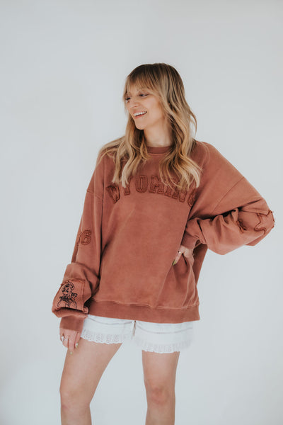 woman smiles and poses with hand on hip in worn, vintage style Wyoming crewneck sweatshirt in brown with stars, Pistol Pete and Wyoming patchwork detailing