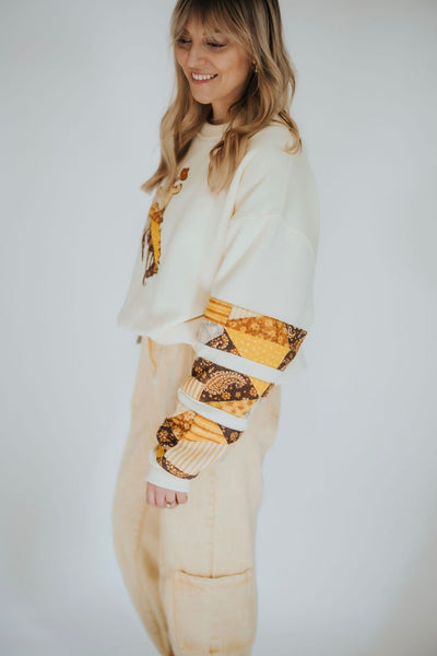 Woman poses in women's University of Wyoming crewneck in cream with the steamboat cowboy and bucking horse on the front and brown and gold patchwork sleeves 