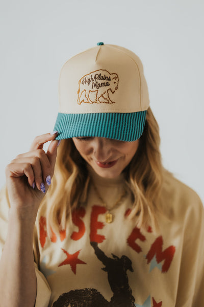 Woman grabs the tip of her snapback turquoise and cream colored hat that reads "high plains mama" with an embroidered buffalo design 
