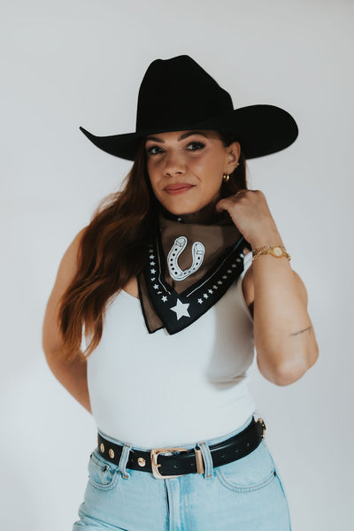Woman wearing a black cowboy hat, white tank top, blue jeans, and a black bandana with star and horseshoe designs.