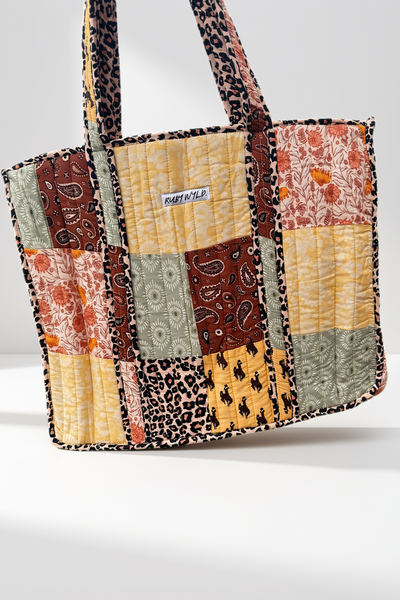 A quilted patchwork tote bag with mixed prints including leopard, paisley, floral, and geometric patterns in yellow, brown, orange, and gray. A small white label reading 'RUBY WYLD"  is visible on the front.